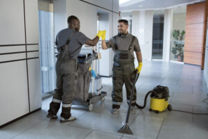 Two janitorial workers wearing safety gloves sharing a fist bump while cleaning a modern office hallway
